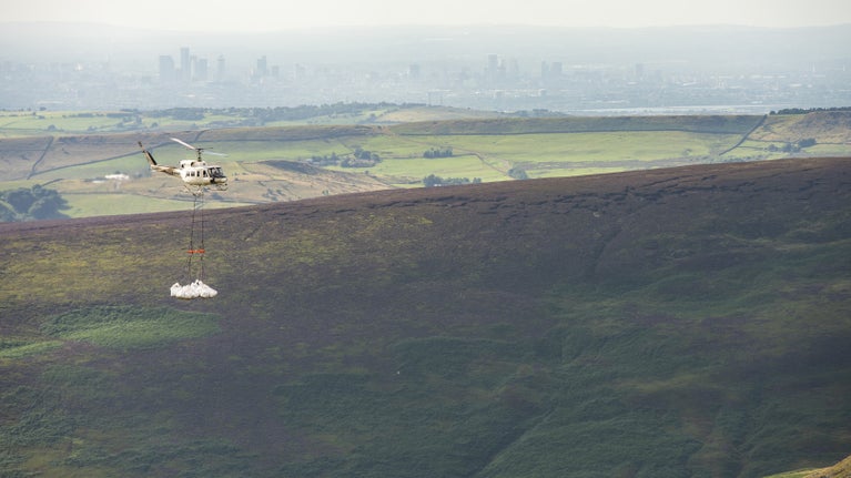 A helicopter flying over moorland on it's way to Kinder Scout to deliver materials for peatland restoration. Manchester can be seen in the background.